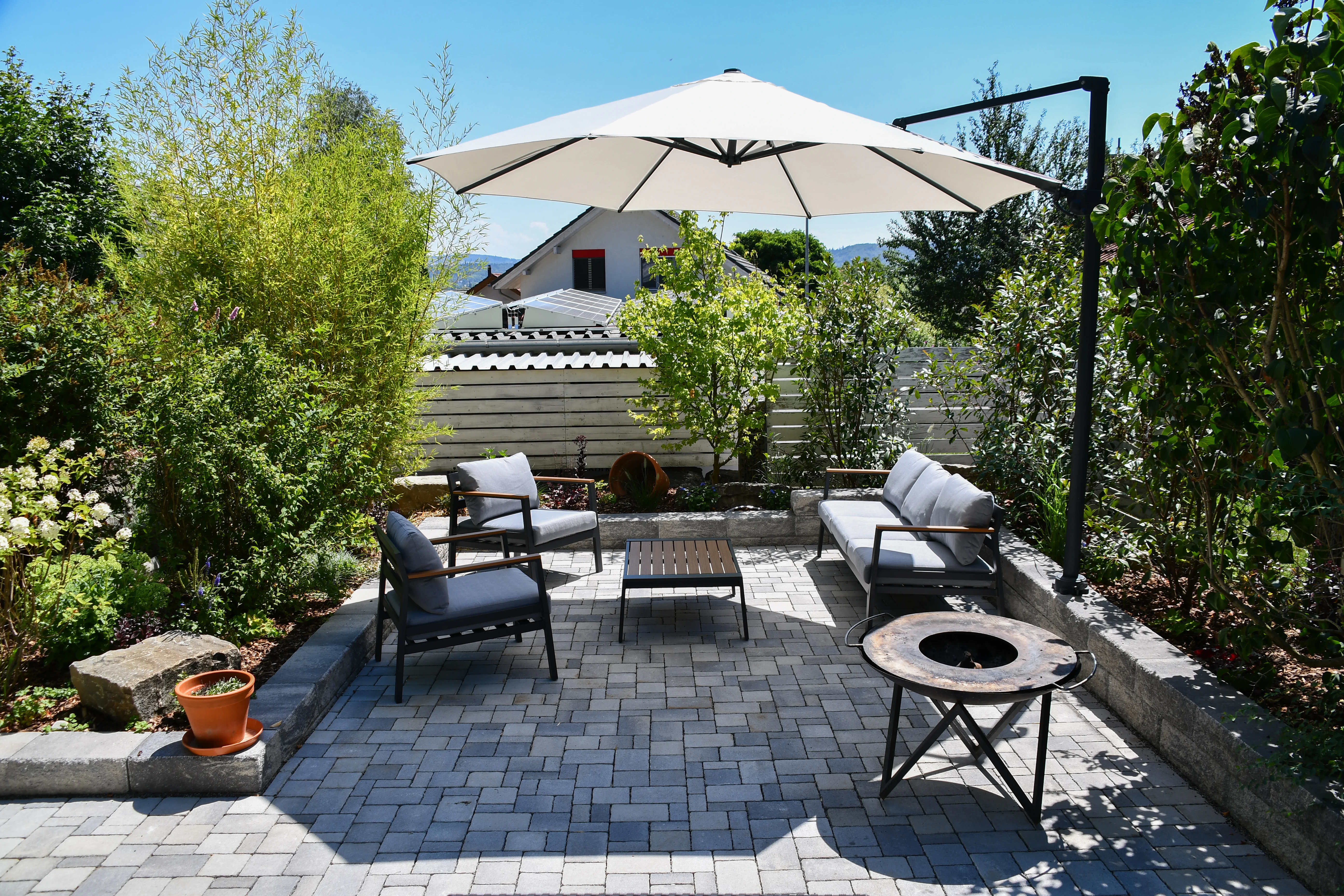 Gartenlounge mit Natursteineinfassung, Pflasterfläche, Sonnenschirm und üppiger Bepflanzung